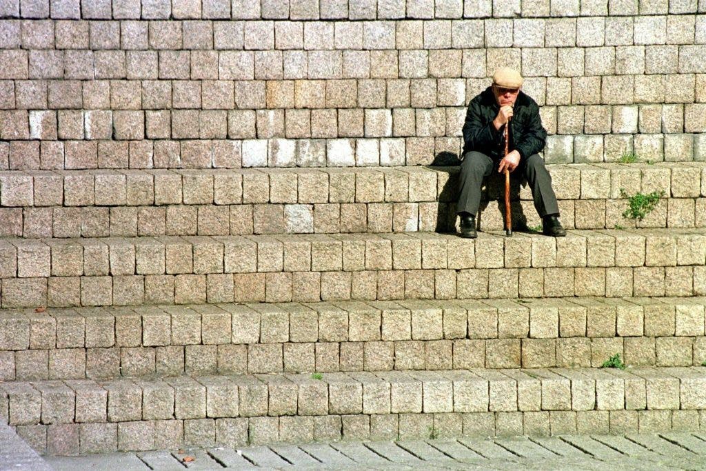 Un anciano, bastón en mano, descansa sentado en unas escaleras de piedra. efesalud.com