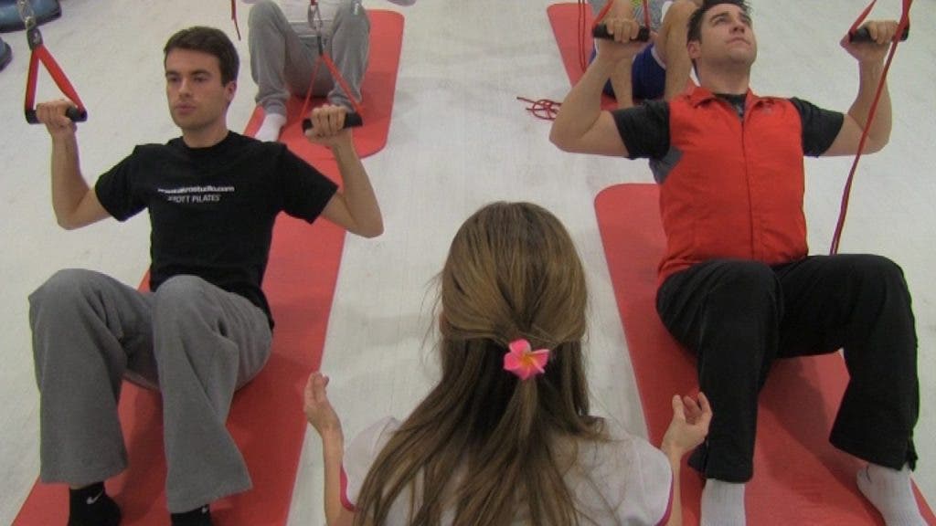 Dos jóvenes, con los glúteos apoyados sobre el suelo del gimnasio, ejercitan su musculación con Redcord, técnica de entrenamiento en suspensión que emplea cuerdas, cinchas y poleas para la sujección.