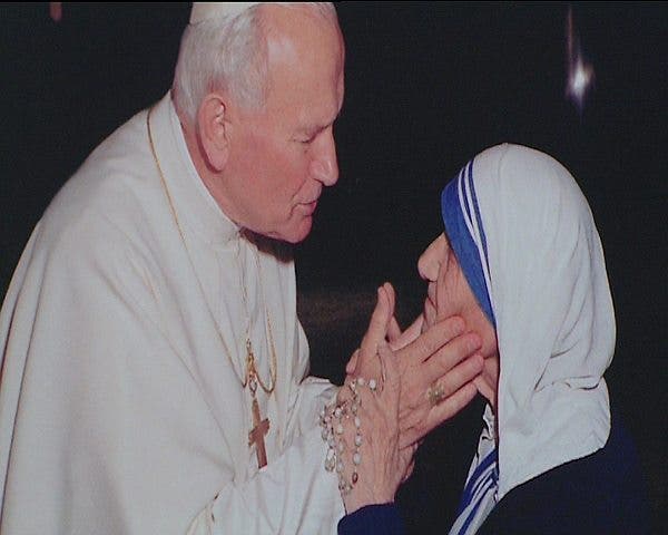 El Papa Juan Pablo II habla con la Madre Teresa de Calcuta, vestidos ambos con sus hábitos habituales, él de blanco con la cruz dorada colgada sobre su pecho y ella con el sari de las Misioneras de la Caridad, blanco y con rayas azules.