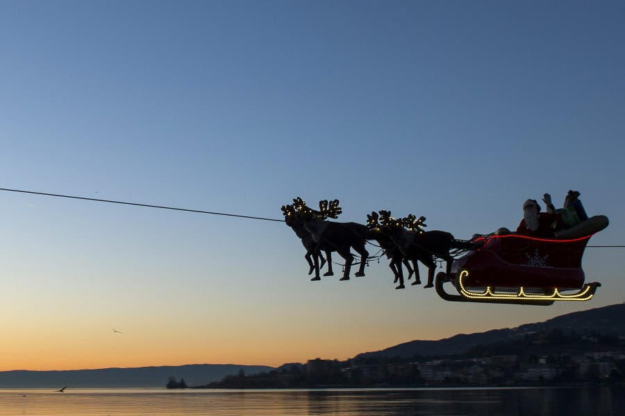 Un hombre vestido de Papá Noel saluda desde su trineo volador conducido por renos desde el Lago de Ginebra frente a los Alpes franceses y suizos al atardecer