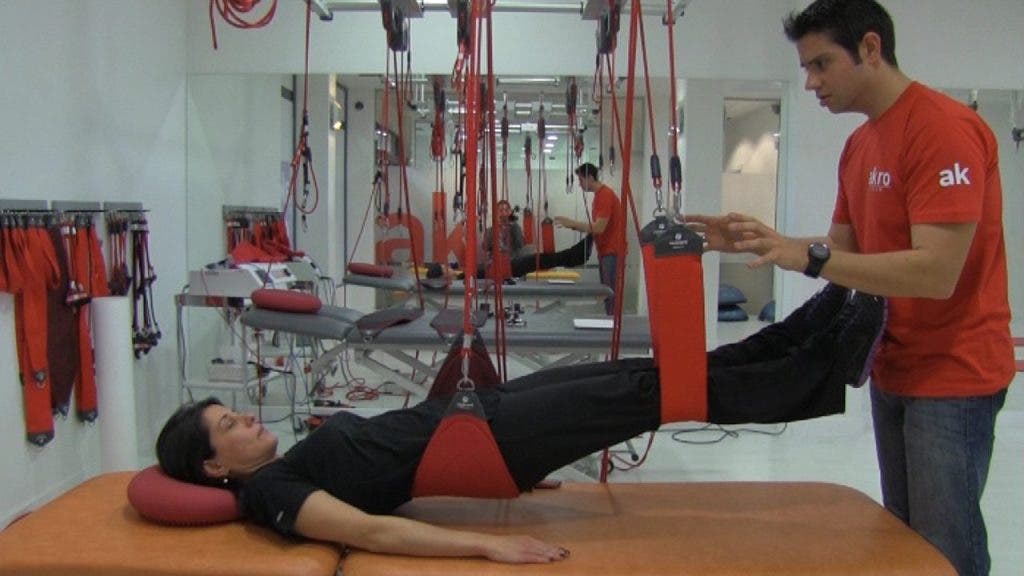 Isabel, tumbada en la camilla con la zona lumbar y sus piernas elevadas por las cuerdas y cinchas de Redcord, trabaja su musculación guiada por el fiisioterapeuta.
