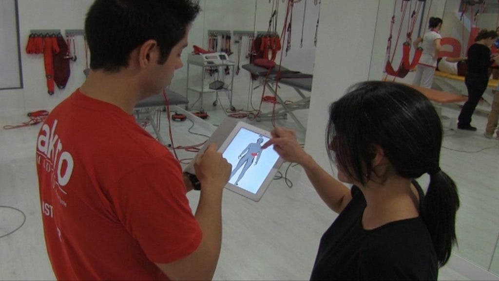 Una mujer, Isabel, señala a Pablo, fisioterapeuta, que sujeta una tableta o ipad con la imagen de una figura humana, la zona lumbar donde tiene un dolor