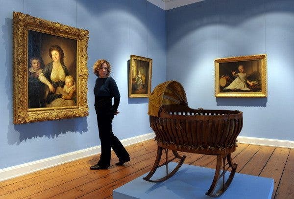 Una mujer, en una exposición de pintura, se vuelve para observar a su espalda una cuna vacia de época. Efesalud.com